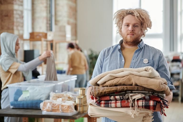A bearded man volunteers indoors, holding donated clothes for charity work amid a collaborative environment.