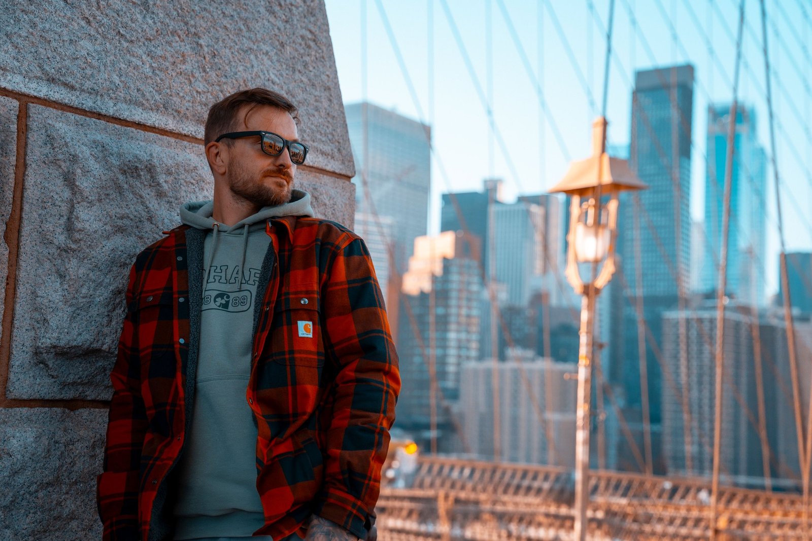 A stylish man in a plaid jacket leaning against the Brooklyn Bridge with the New York City skyline in the background.