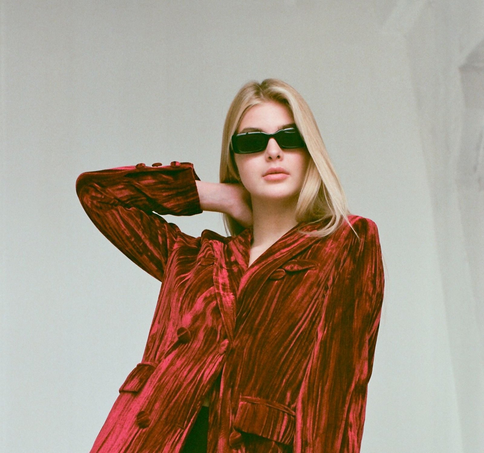 Fashionable woman in red velvet jacket posing indoors with sunglasses.